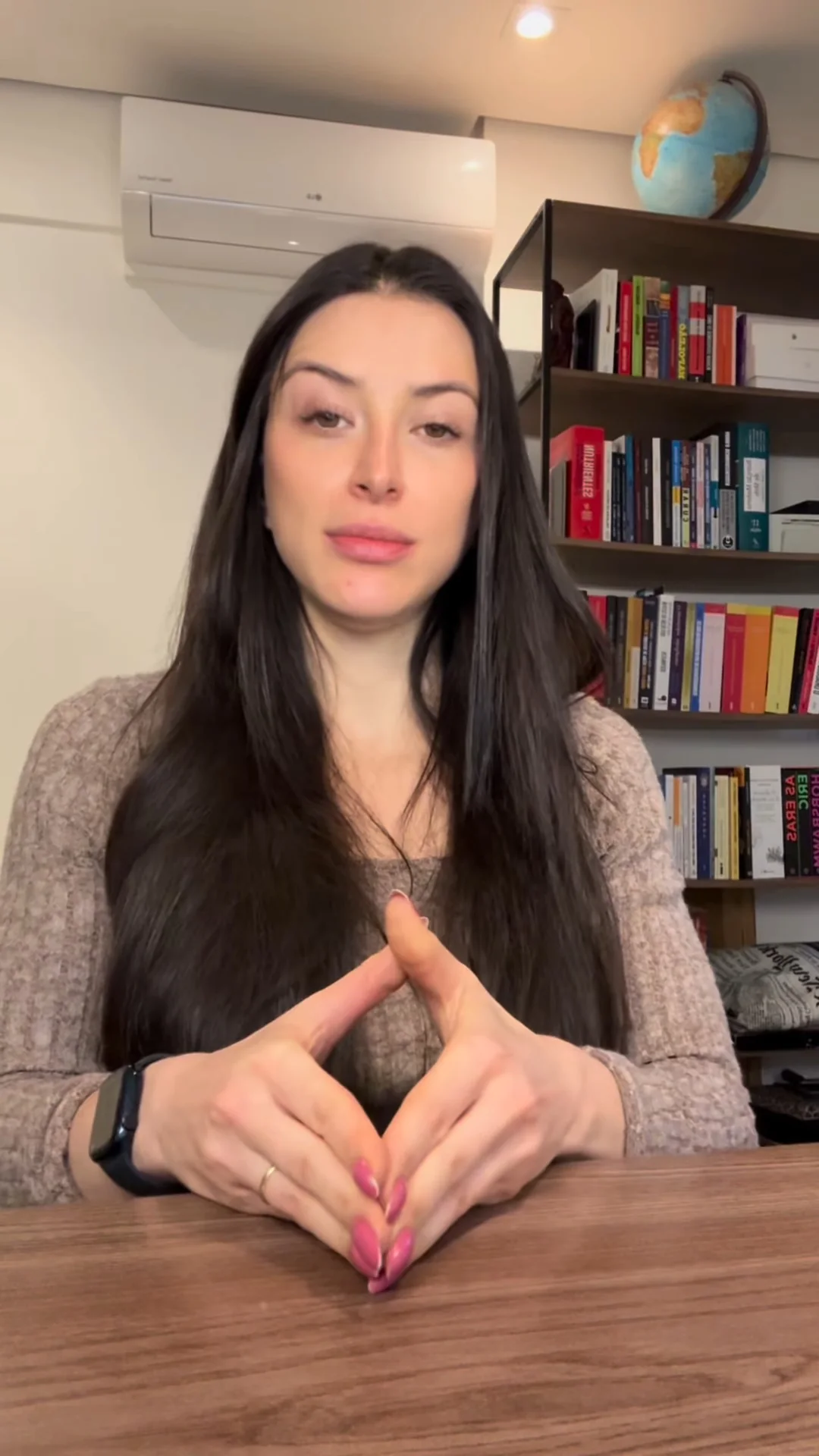 Deborah Moss sentada à mesa de madeira, de blusa de tricô, com as mãos unidas em frente a uma estante de livros
