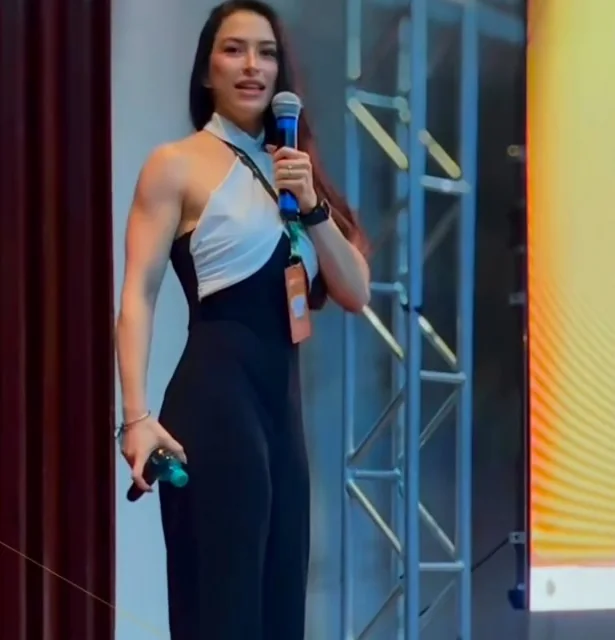 Deborah Moss no palco de um evento em Goiânia, de macacão preto e blusa branca, falando ao microfone