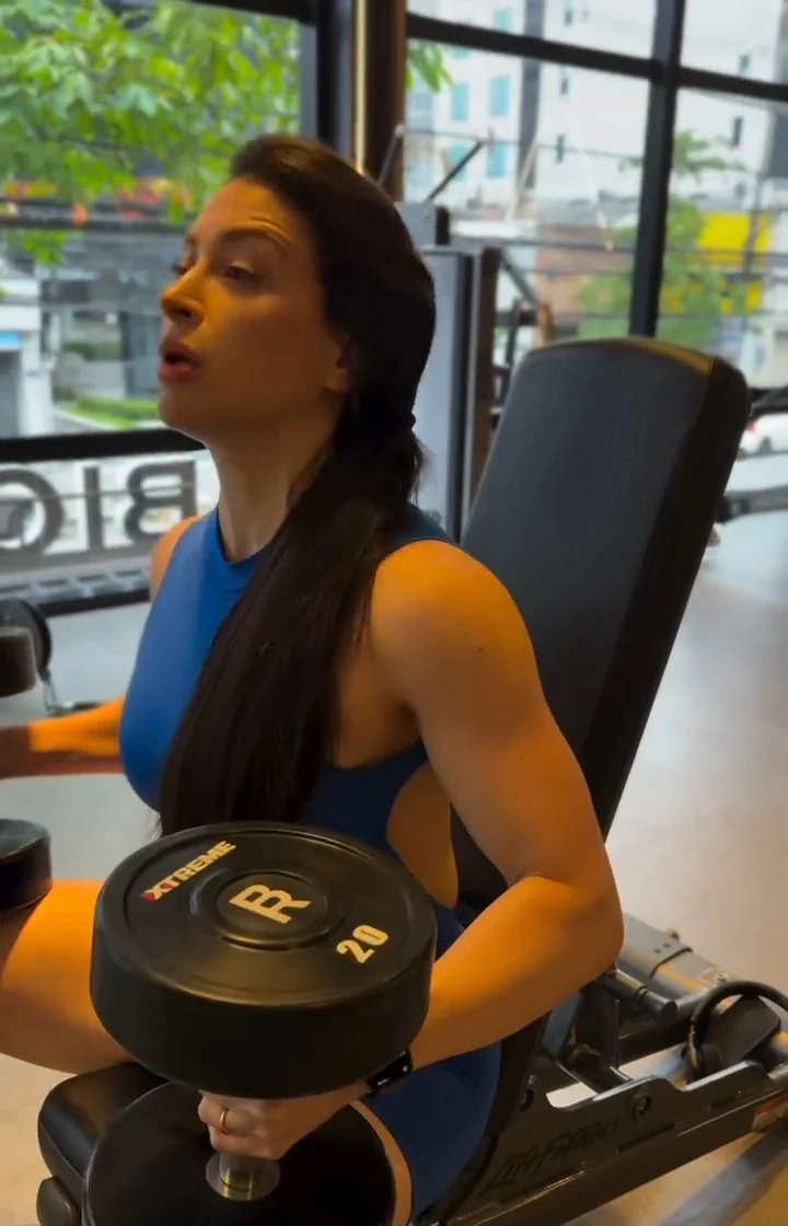 Deborah Moss sentada em um banco de academia, de macaquinho azul, segurando um halter de 20 quilos com concentração