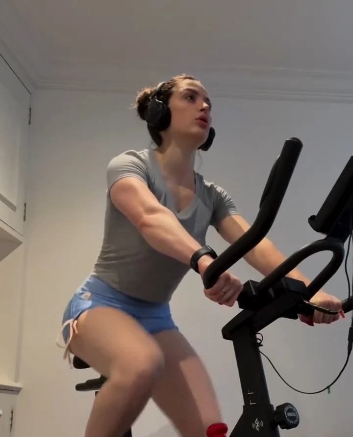 Deborah Moss pedalando em pé em uma bicicleta ergométrica, de fone de ouvido, em um sprint de intensidade alta